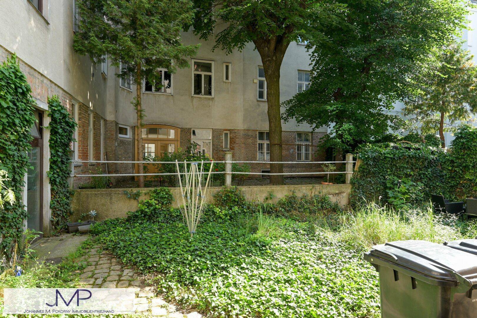 Hinterhofgarten mit viel Grün, Bäumen und einem gepflasterten Bereich, umgeben von Gebäuden.