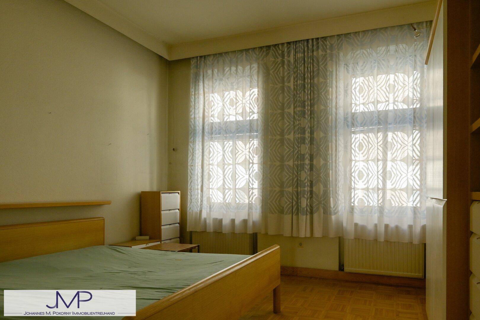 Schlafzimmer mit Parkettboden, Bett und zwei Fenstern mit Vorhängen, bietet viel Tageslicht.