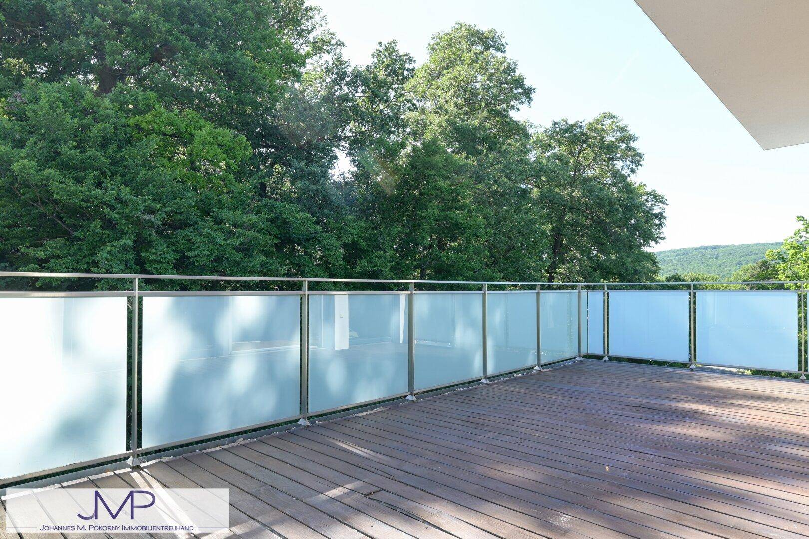 Geräumiger Balkon mit Holzdeck und modernem Glasgeländer, umgeben von üppigem Grün.