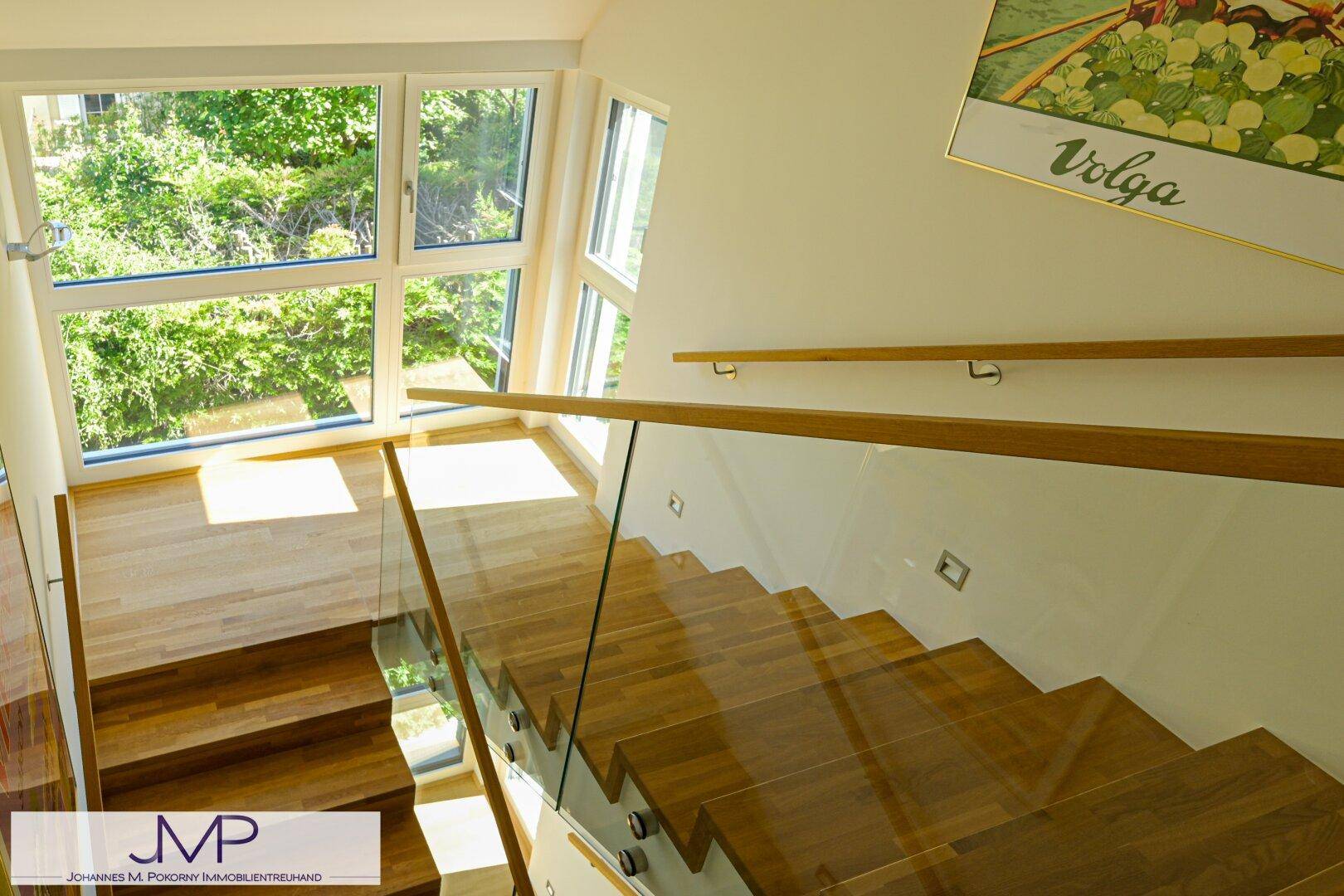 Helle Holztreppe mit Glasgeländer und großen Fenstern, die viel natürliches Licht hereinlassen.