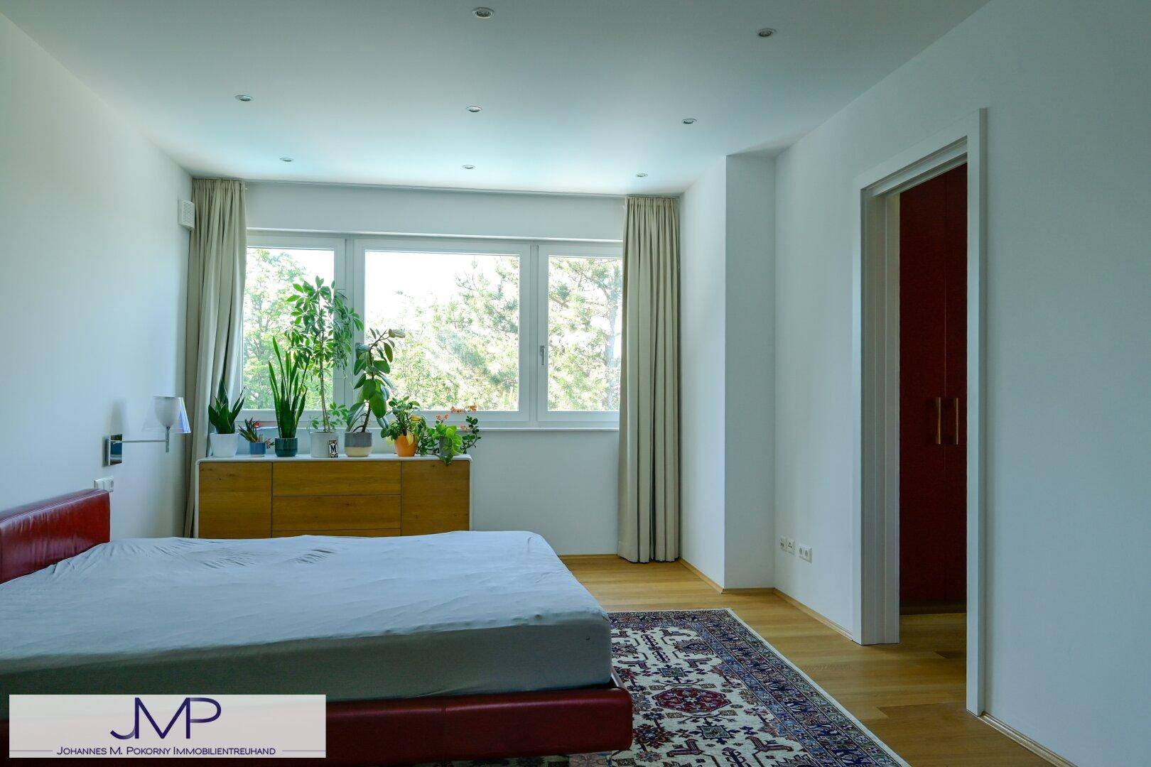 Helles Schlafzimmer mit rotem Bett, Holzkommode und großem Fenster mit Blick ins Grüne.