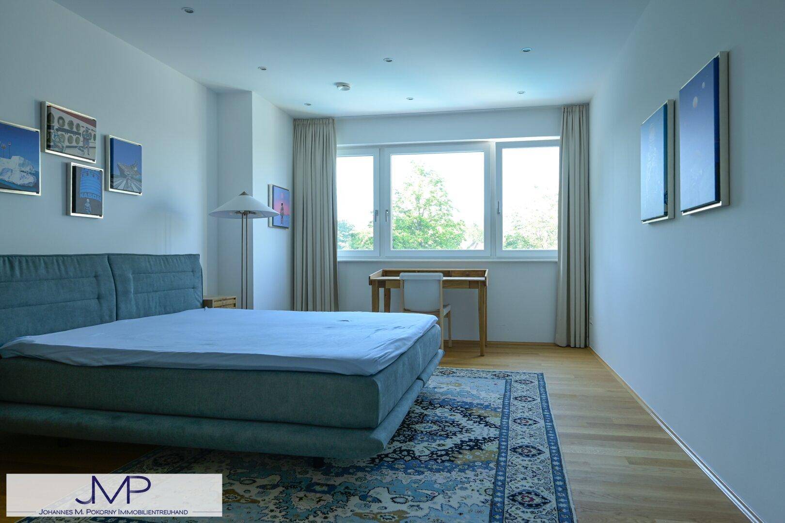 Helles Schlafzimmer mit blauem Bett, Schreibtisch am Fenster und dekorativen Bildern an den Wänden.