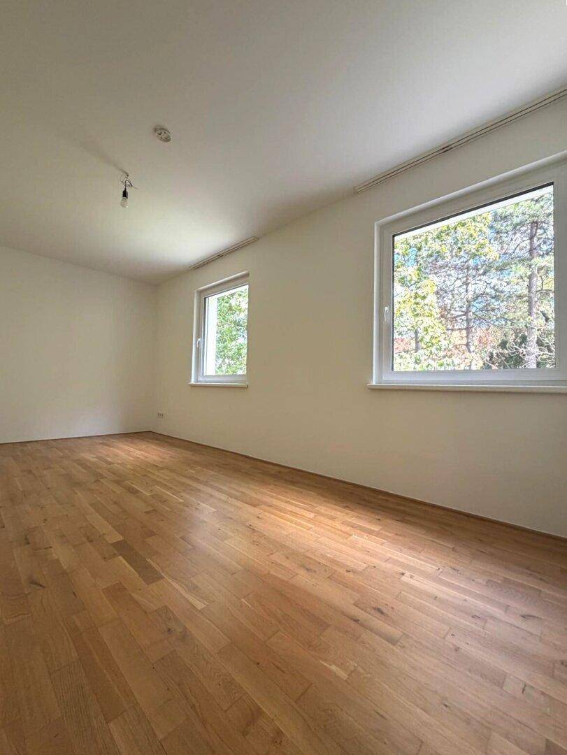 Leeres Zimmer mit Parkettboden und großen Fenstern, die viel Licht hereinlassen.