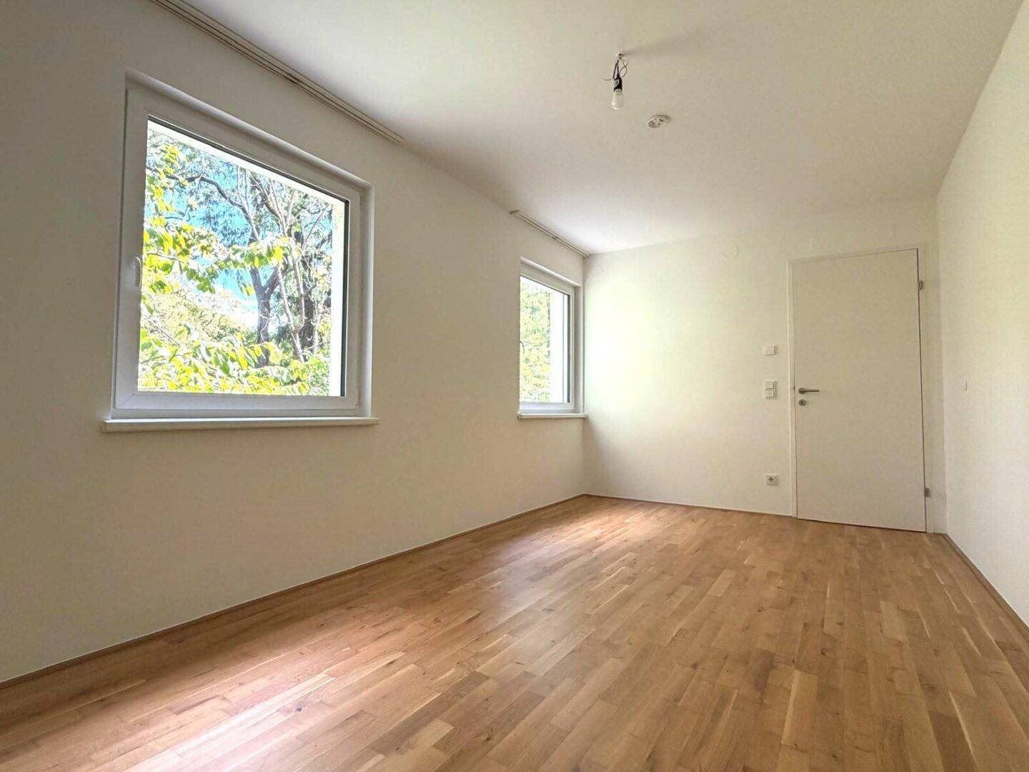 Helles Schlafzimmer mit Holzboden und zwei Fenstern mit Blick ins Grüne.
