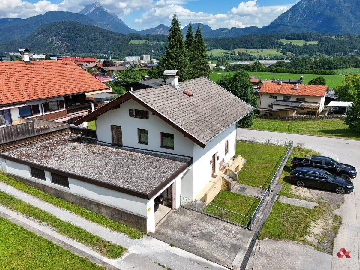 Luftaufnahme des Hauses mit Flachdachgarage, Garten und umliegender Wohnbebauung vor Bergkulisse.
