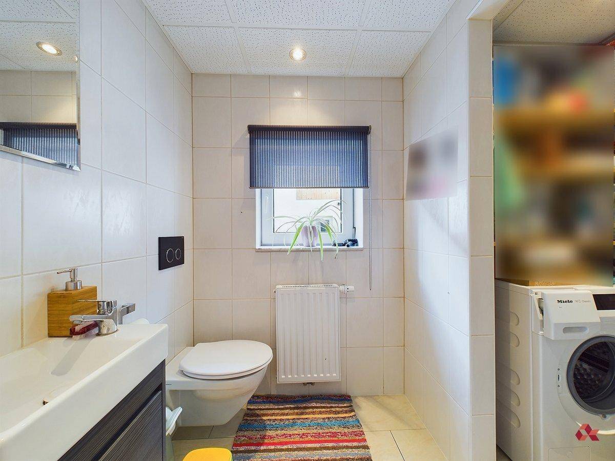 Kleines WC mit weißen Fliesen, Waschbecken und Waschmaschine, gut beleuchtet durch ein Fenster.