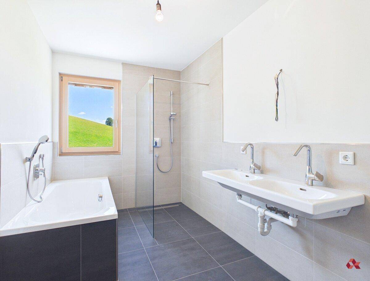 Modernes Badezimmer mit Badewanne, Dusche, Doppelwaschbecken und Fenster mit Grünblick.
