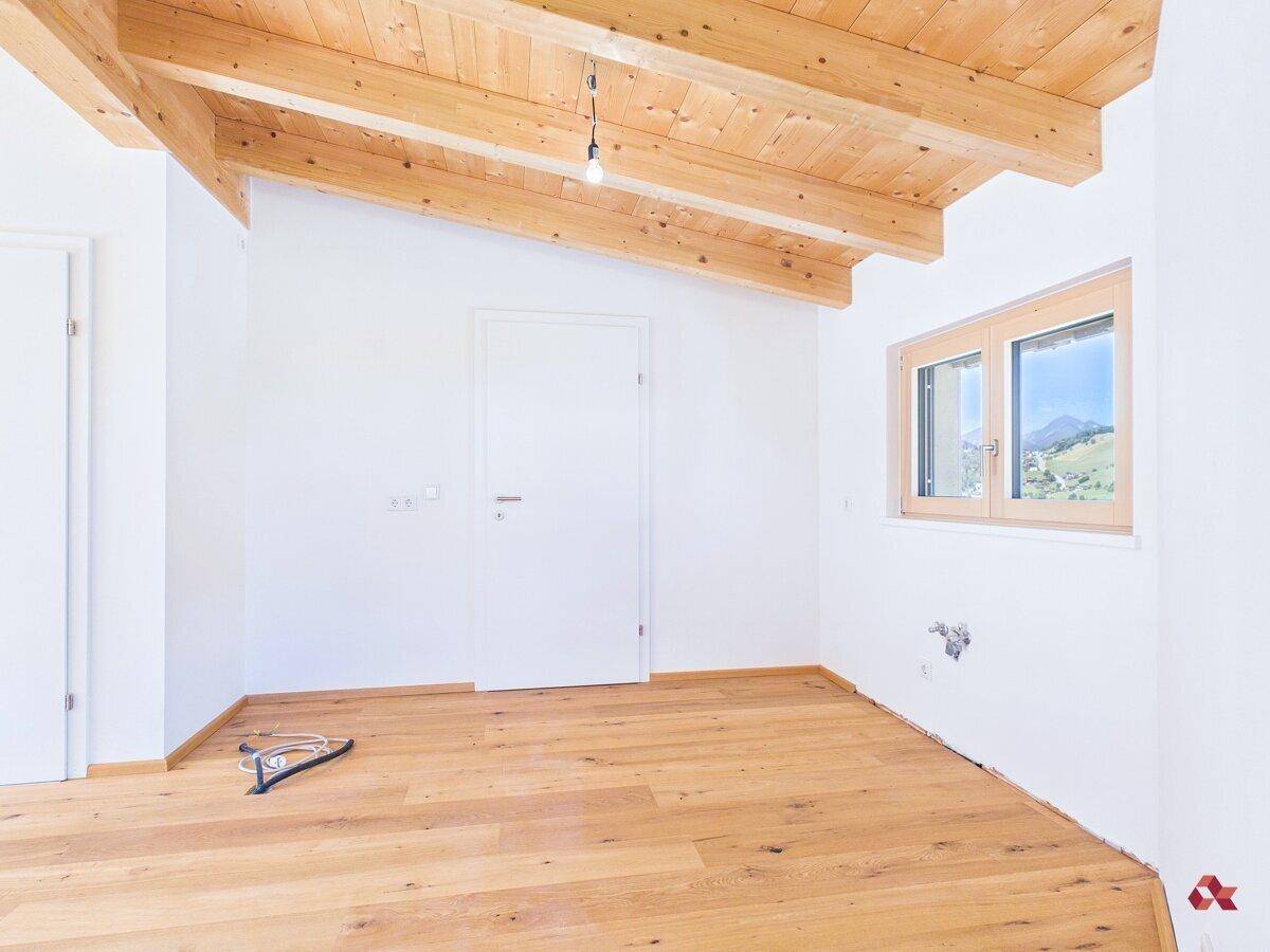 Helles, leeres Zimmer mit Holzbalkendecke, Parkettboden und Fenster mit Bergblick.