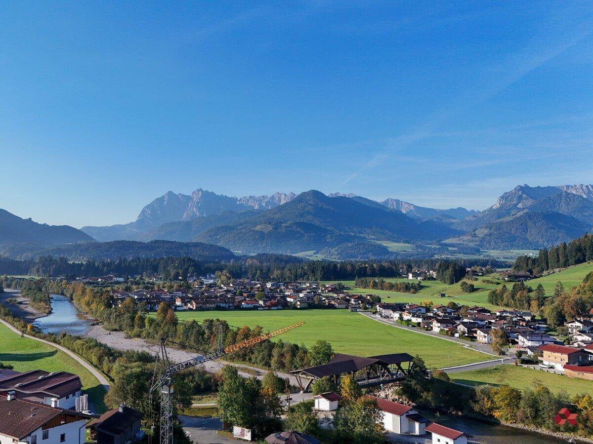 Malerische Landschaft mit Fluss, Dorf und imposantem Bergpanorama unter blauem Himmel.