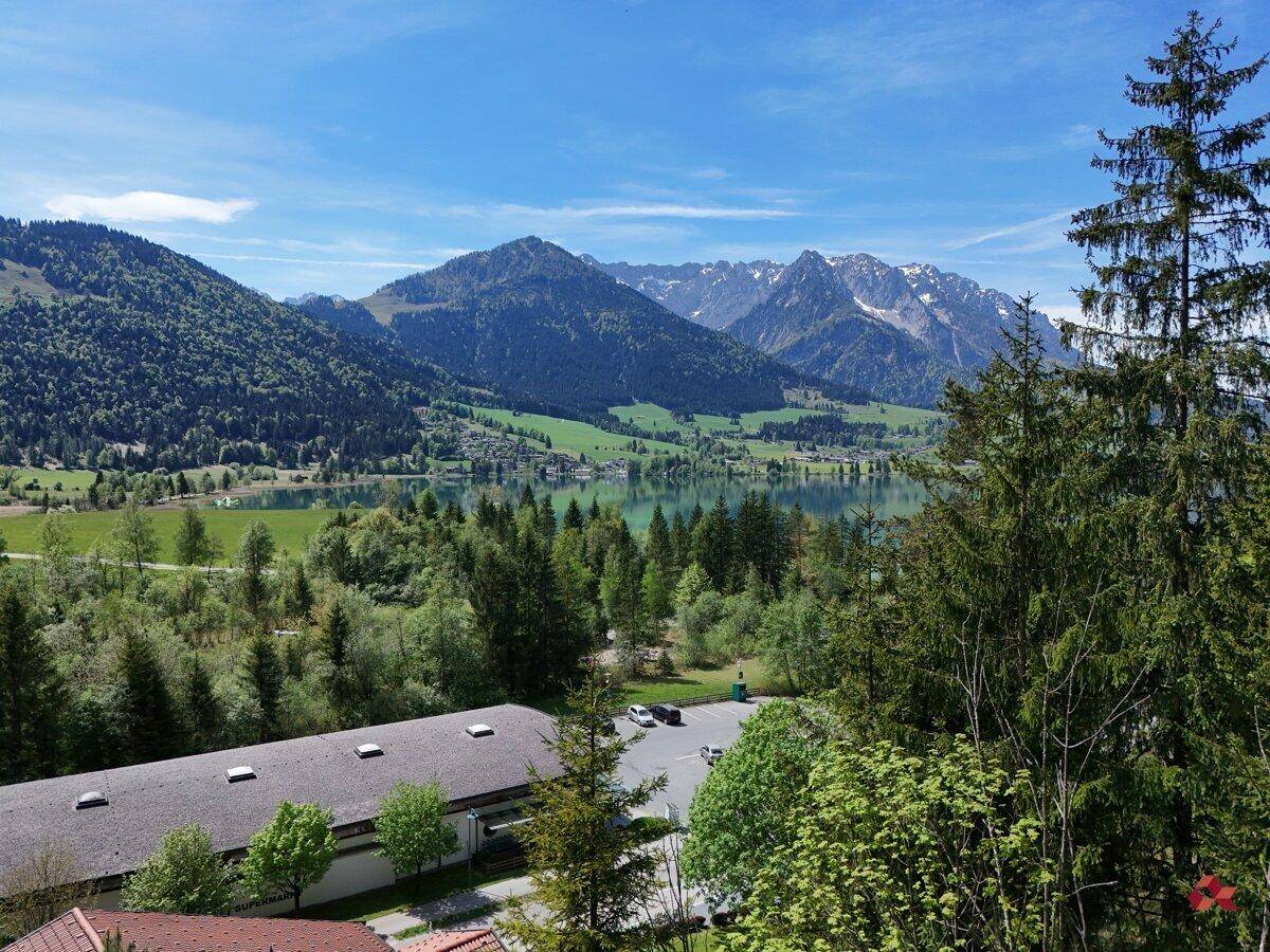 Weitläufiger Ausblick auf einen türkisfarbenen See, umrahmt von bewaldeten Bergen und einer klaren Himmelslandschaft.