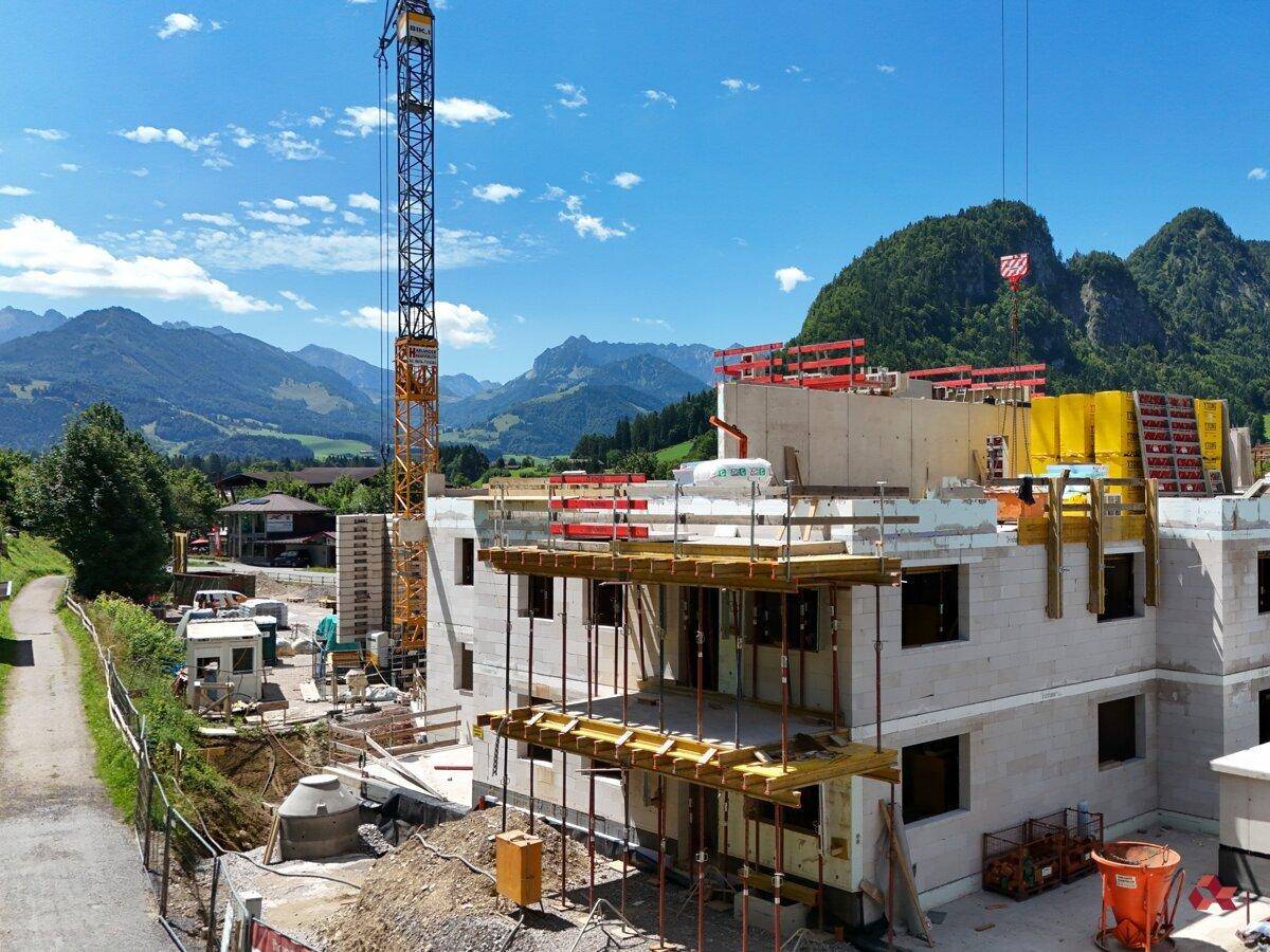 Baustelle eines Neubaus mit sichtbarer Betonstruktur und Kran, umgeben von grünen Hügeln und Bergen.