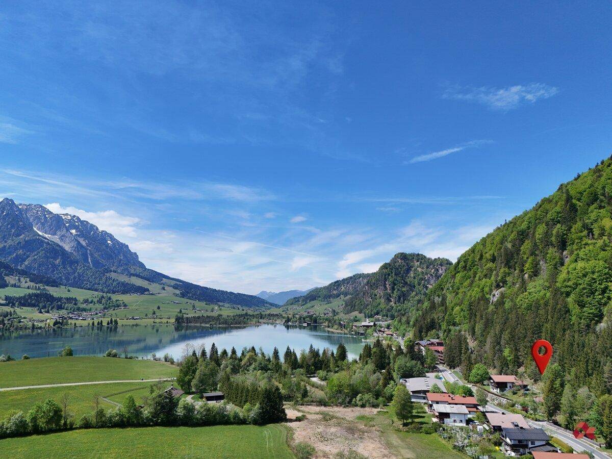 Weitwinkelaufnahme eines idyllischen Sees mit umliegenden Bergen und einem Dorf, markiert mit einem Standort-Pin.