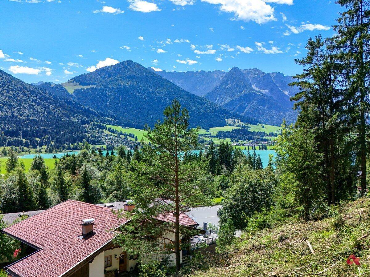 Malerische Landschaft mit einem traditionellen Haus, umgeben von Bergen, Wald und einem klaren See unter blauem Himmel.