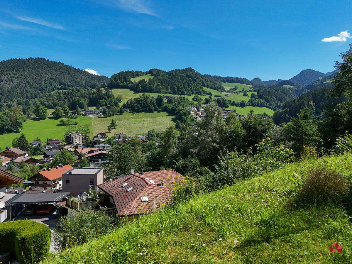 Panoramablick auf eine idyllische Siedlung in einem grünen Tal, umgeben von bewaldeten Hügeln unter blauem Himmel.