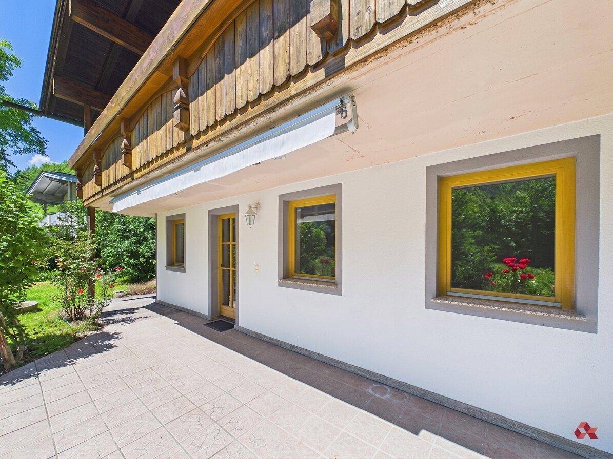 Helle Terrasse mit Fliesenboden und Zugang zum Haus, umgeben von grüner Bepflanzung.