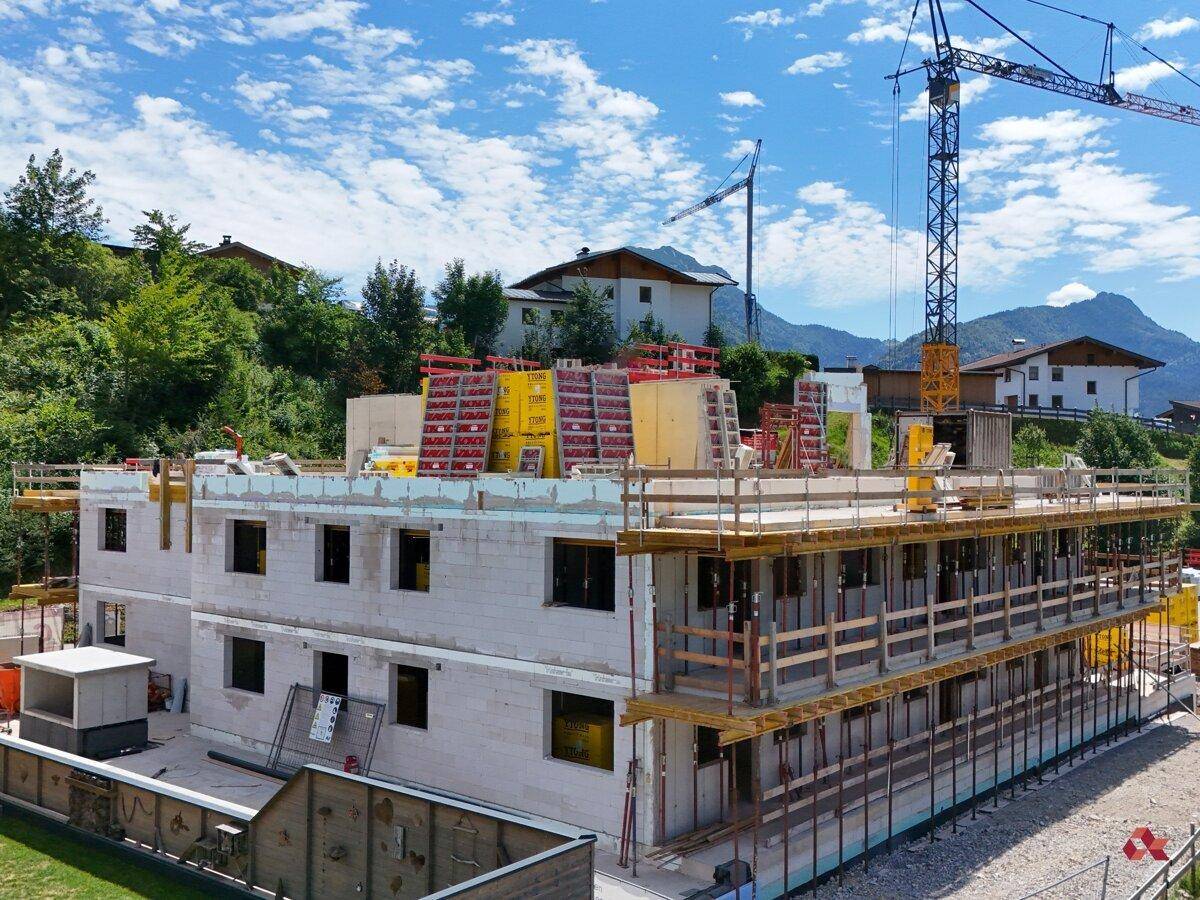 Baustelle eines mehrstöckigen Gebäudes mit Gerüst und Kran, vor einer Kulisse aus Häusern und Bergen.