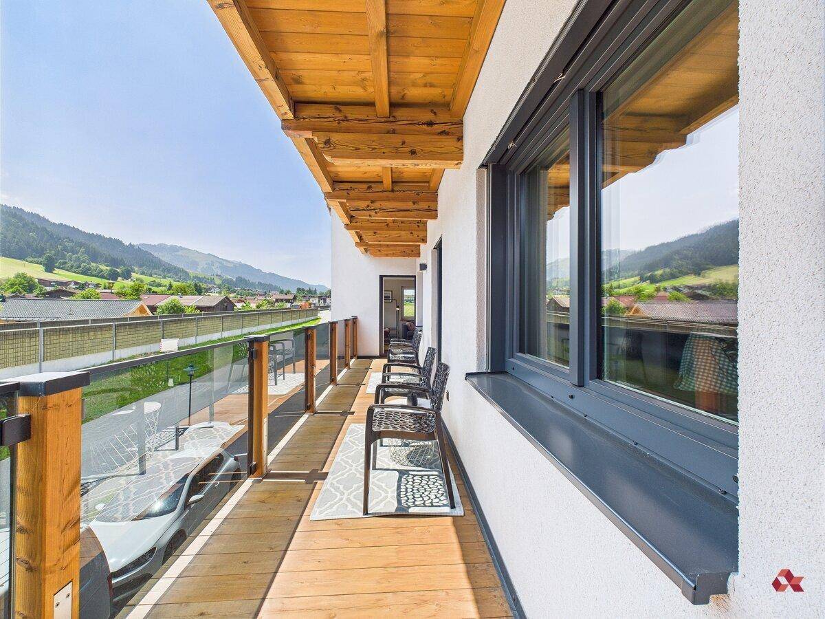 Langer Balkon mit Holzboden, Glasgeländer und Sitzgelegenheiten, bietet weite Aussicht auf die Berge.