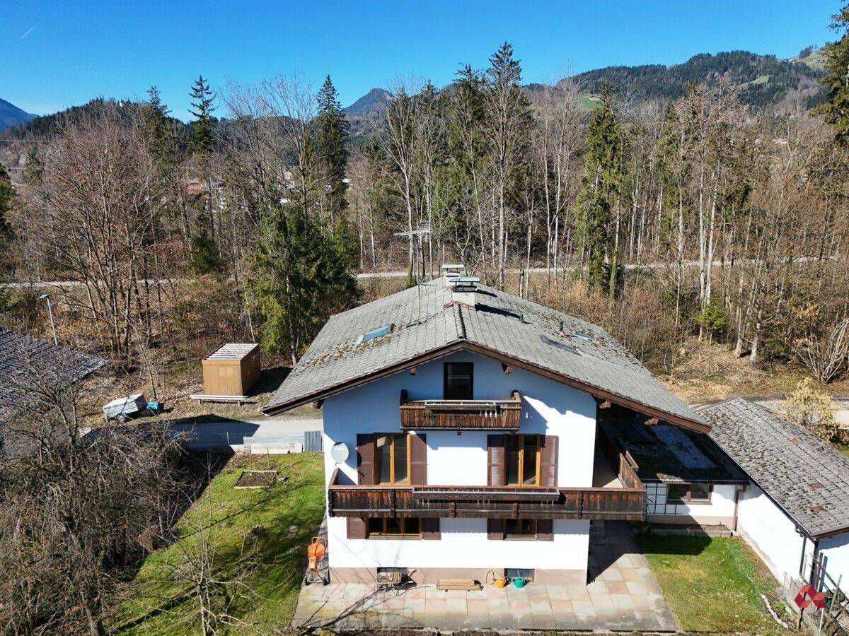 Luftaufnahme des Hauses mit Satteldach und Balkonen, umgeben von Bäumen und Bergen.