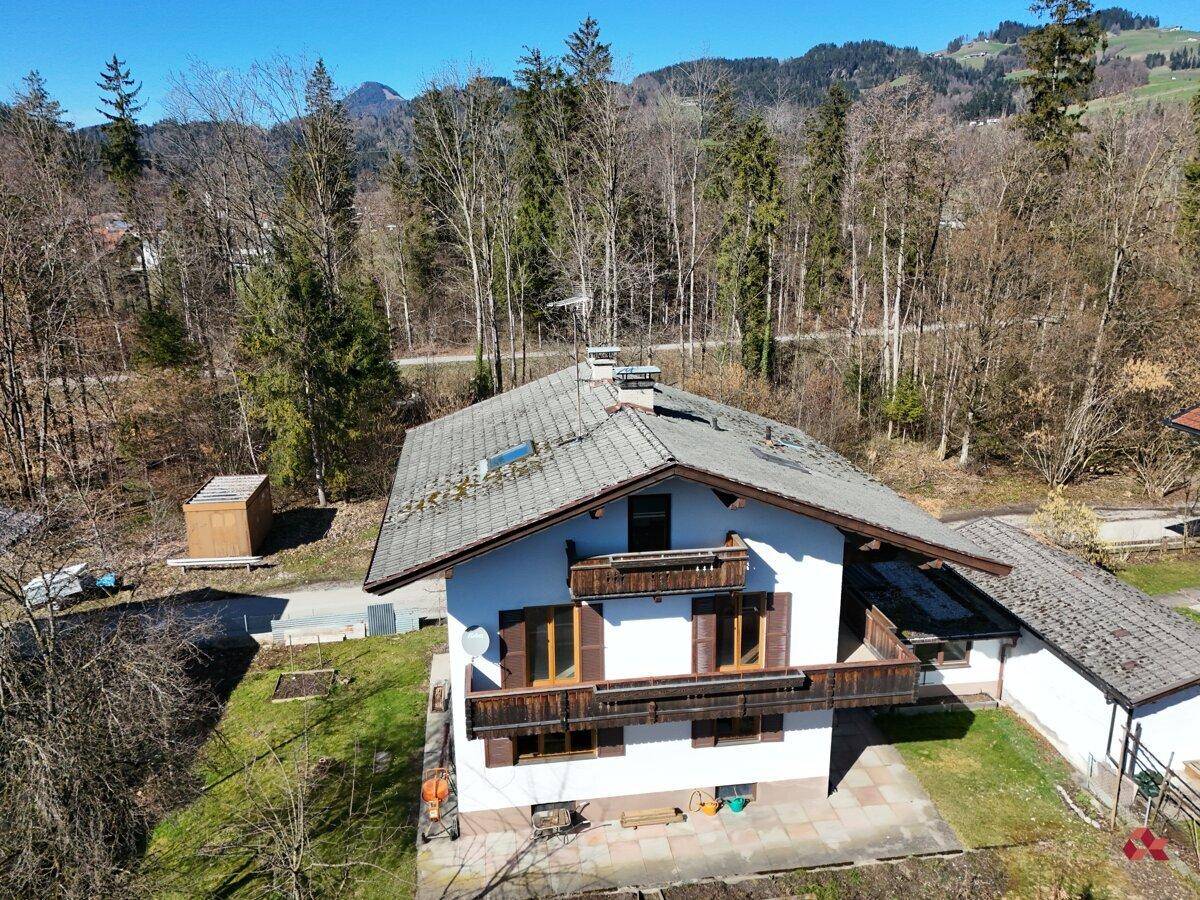 Luftaufnahme des Hauses mit Satteldach und Balkonen, umgeben von Bäumen und Bergen.