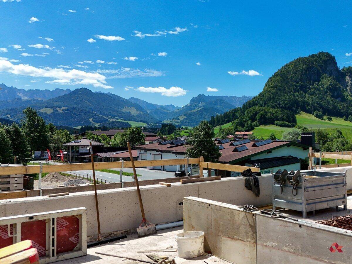 Blick von der Baustelle auf die umliegende Berglandschaft und grüne Wiesen unter blauem Himmel.