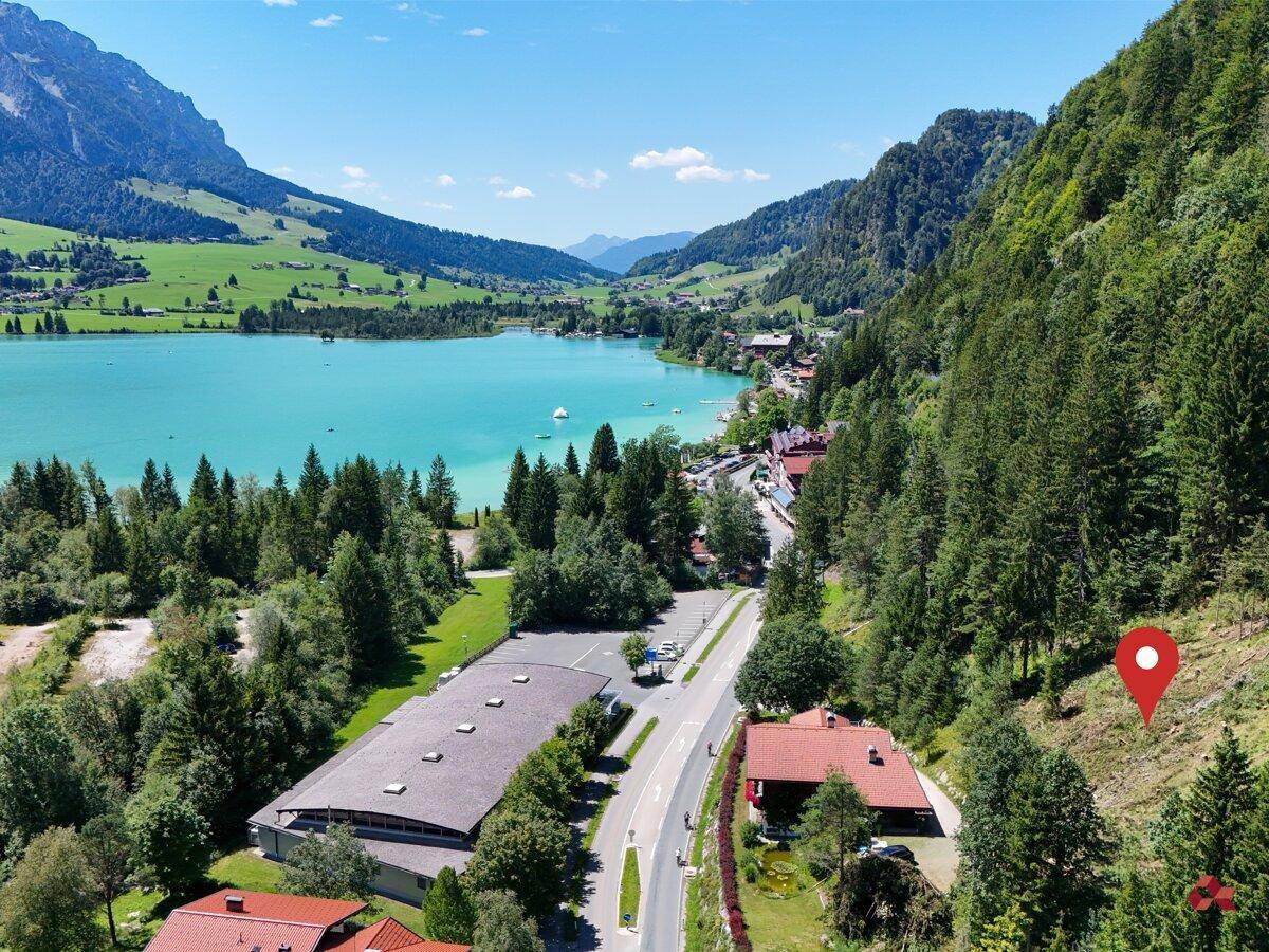 Luftaufnahme einer Siedlung am Ufer eines türkisfarbenen Sees, umgeben von bewaldeten Bergen.