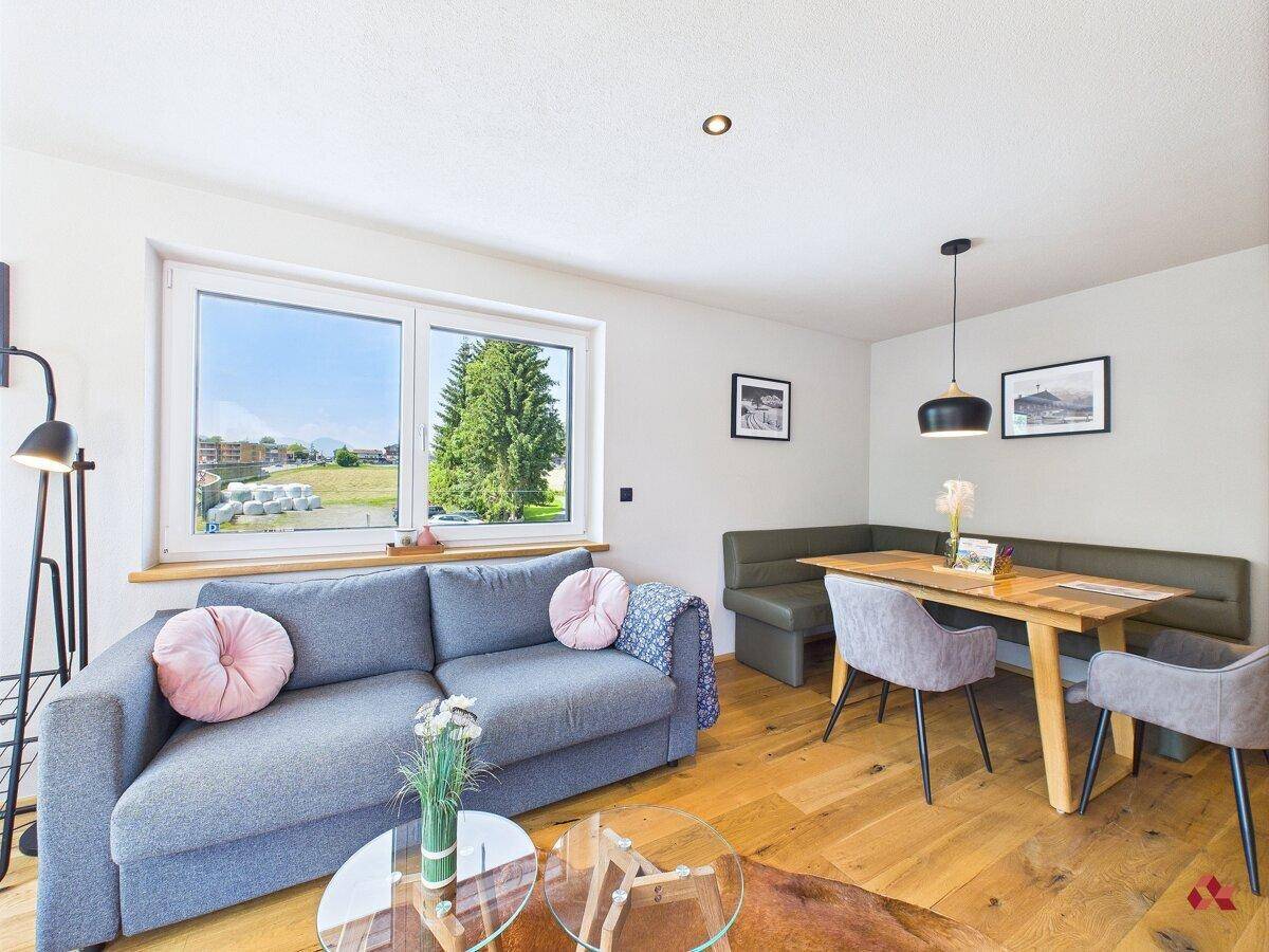 Wohnbereich mit Sofa und Esstisch, Holzboden und großem Fenster mit Blick ins Grüne.