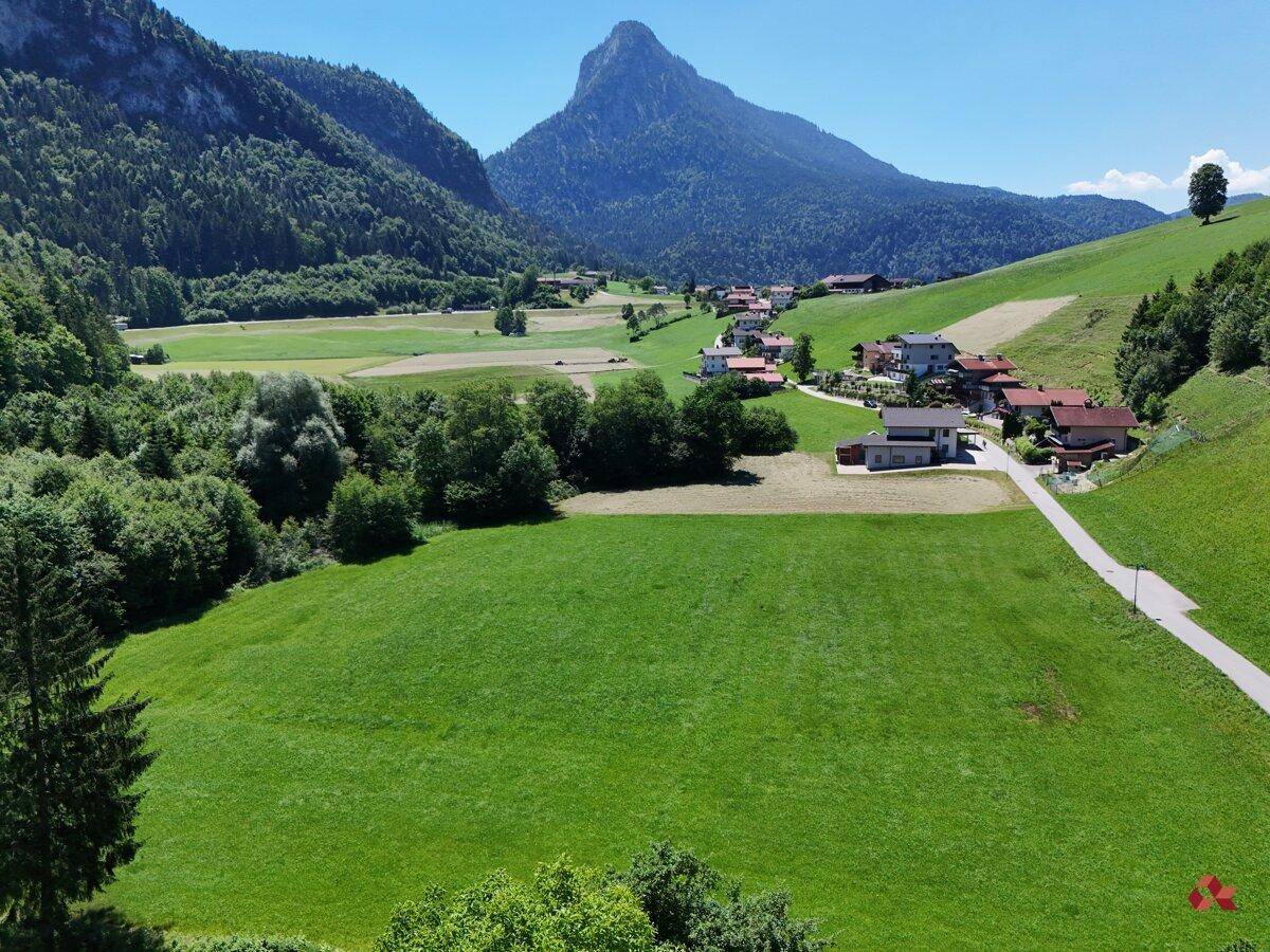 Weitläufige grüne Landschaft mit Bergen im Hintergrund und einigen Häusern in einer ländlichen Umgebung.