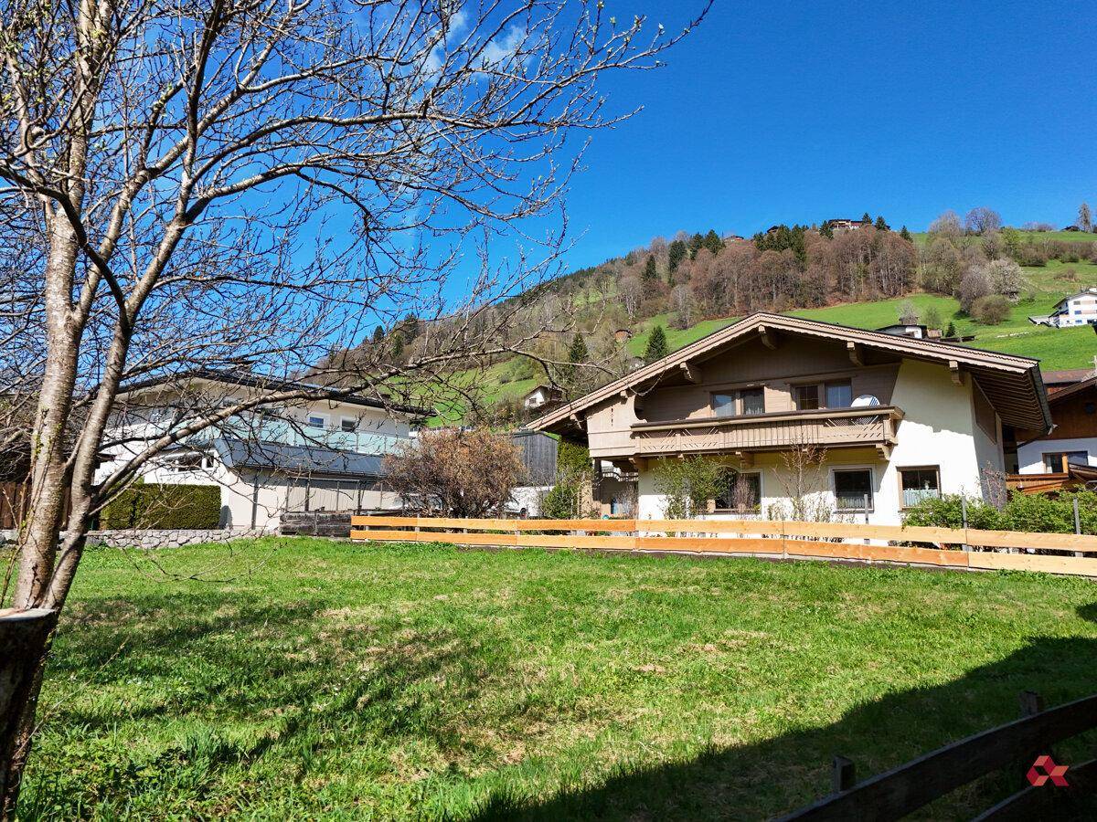 Traditionelles Haus in den Bergen mit grüner Wiese und blühendem Baum im Vordergrund unter blauem Himmel.