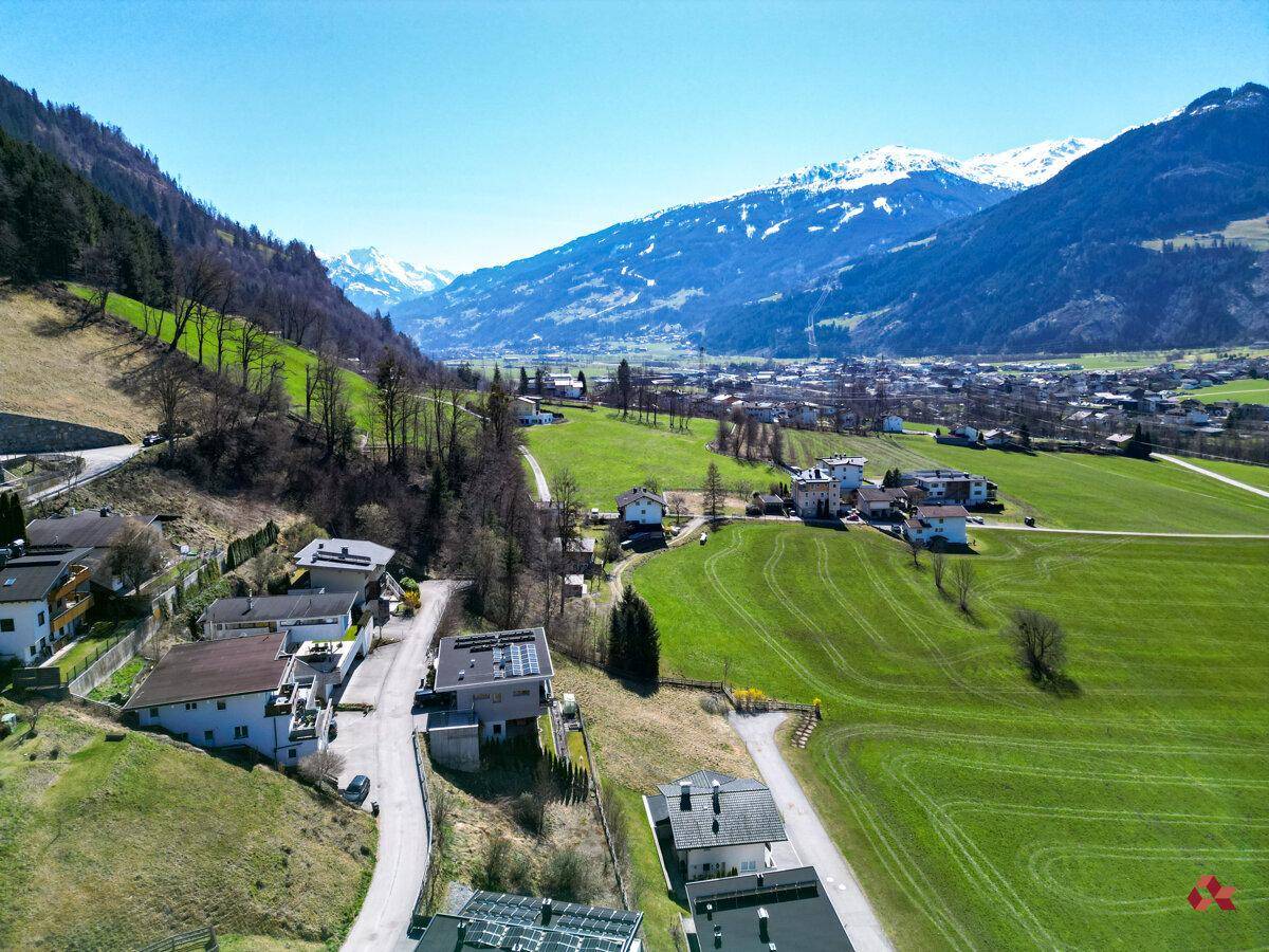 Luftaufnahme einer Siedlung mit Häusern und Straßen, umgeben von grünen Feldern und Bergen.