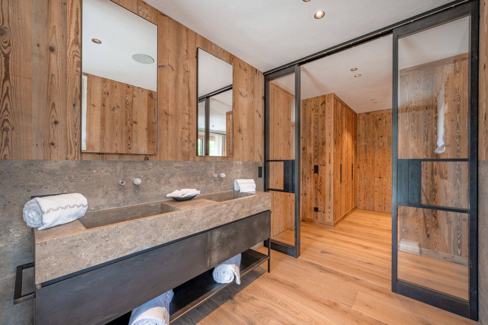 Modernes Badezimmer mit Doppelwaschbecken aus Stein und Holzwänden, verbunden mit einem Ankleidebereich.