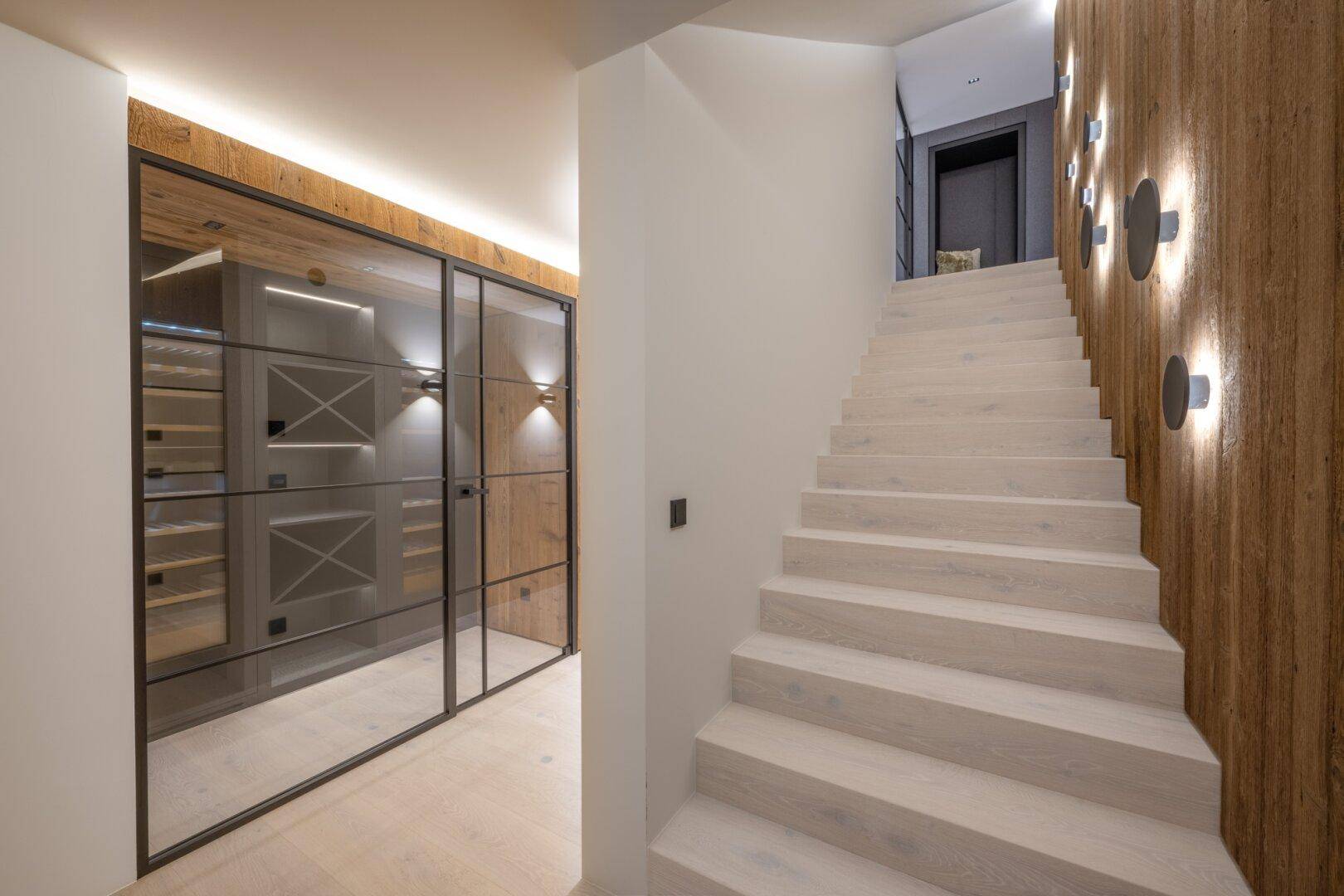 Heller Flur mit moderner Treppe und einem beeindruckenden, verglasten Weinkeller als Blickfang.