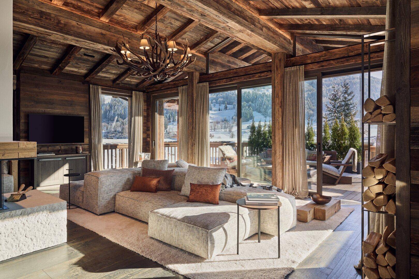 Helles Wohnzimmer mit großem Sofa, Hirschgeweih-Kronleuchter und Zugang zur verschneiten Terrasse mit Bergblick.