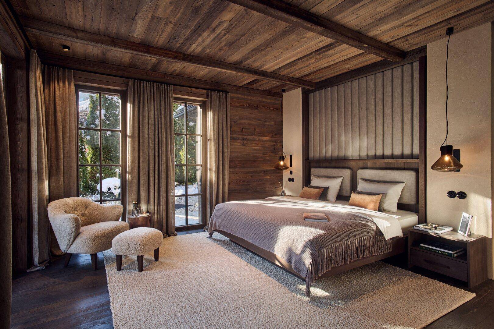 Gemütliches Schlafzimmer mit großem Bett, Holzwänden und -decken sowie großen Fenstern mit Blick ins Grüne.