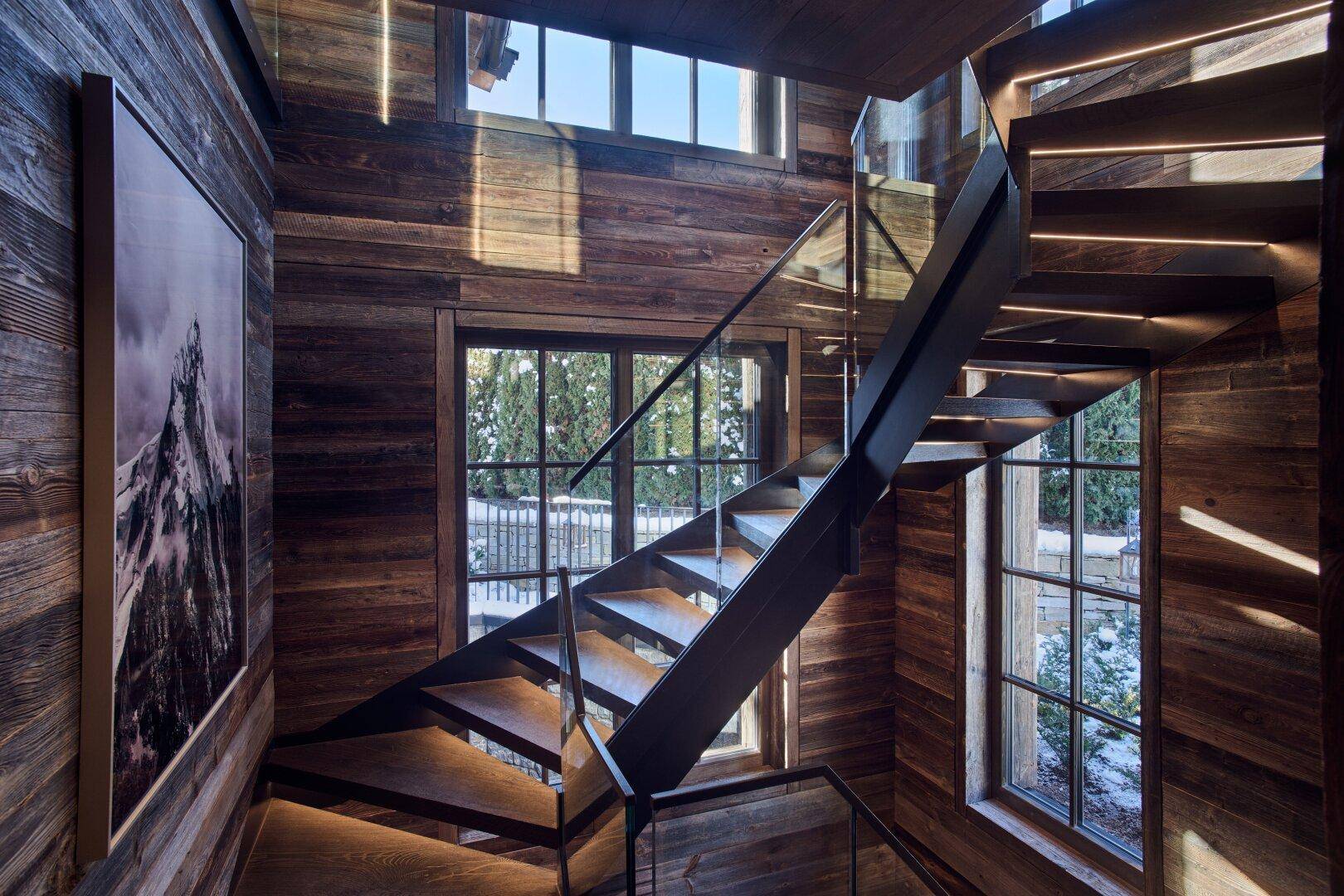 Moderne Holztreppe mit Glasgeländer und integrierter Beleuchtung, die das natürliche Licht der Fenster einfängt.
