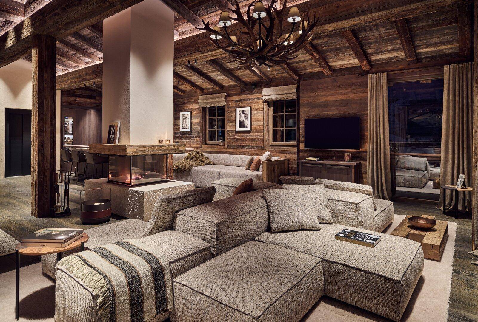 Geräumiges Wohnzimmer mit großem Sofa, Kamin, Holzbalken und einem Hirschgeweih-Kronleuchter.