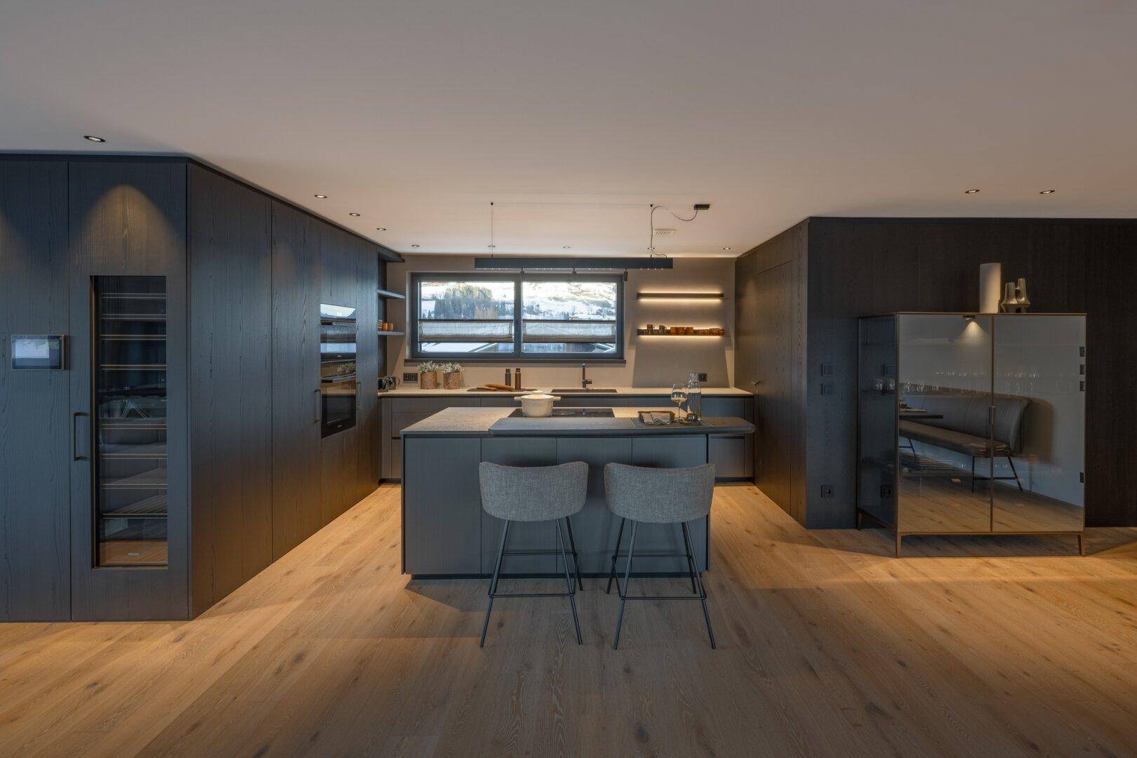 Elegante, moderne Küche in dunklen Tönen mit Kücheninsel, integrierten Geräten und Fensterblick.
