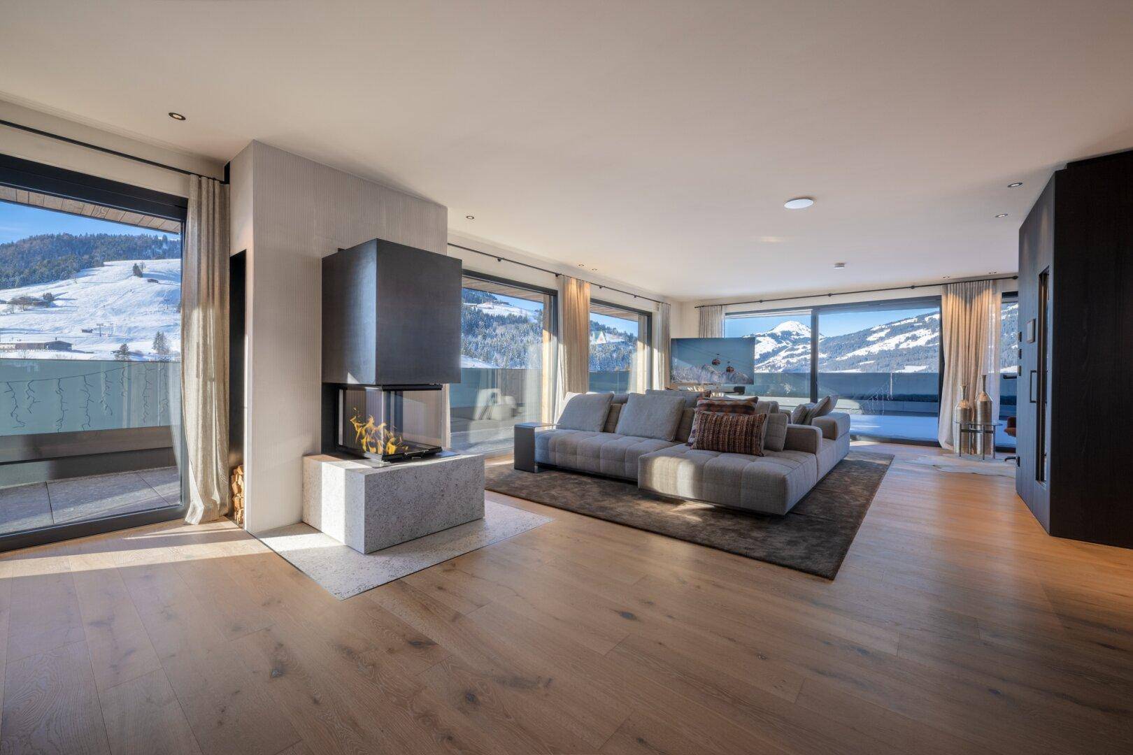 Offener Wohnbereich mit modernem Kamin, Holzboden und weitem Blick auf die winterliche Berglandschaft.