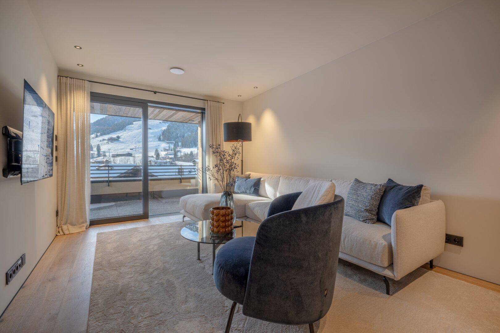 Helles Wohnzimmer mit bequemer Sitzecke, Fernseher und Zugang zu einem Balkon mit Bergblick.