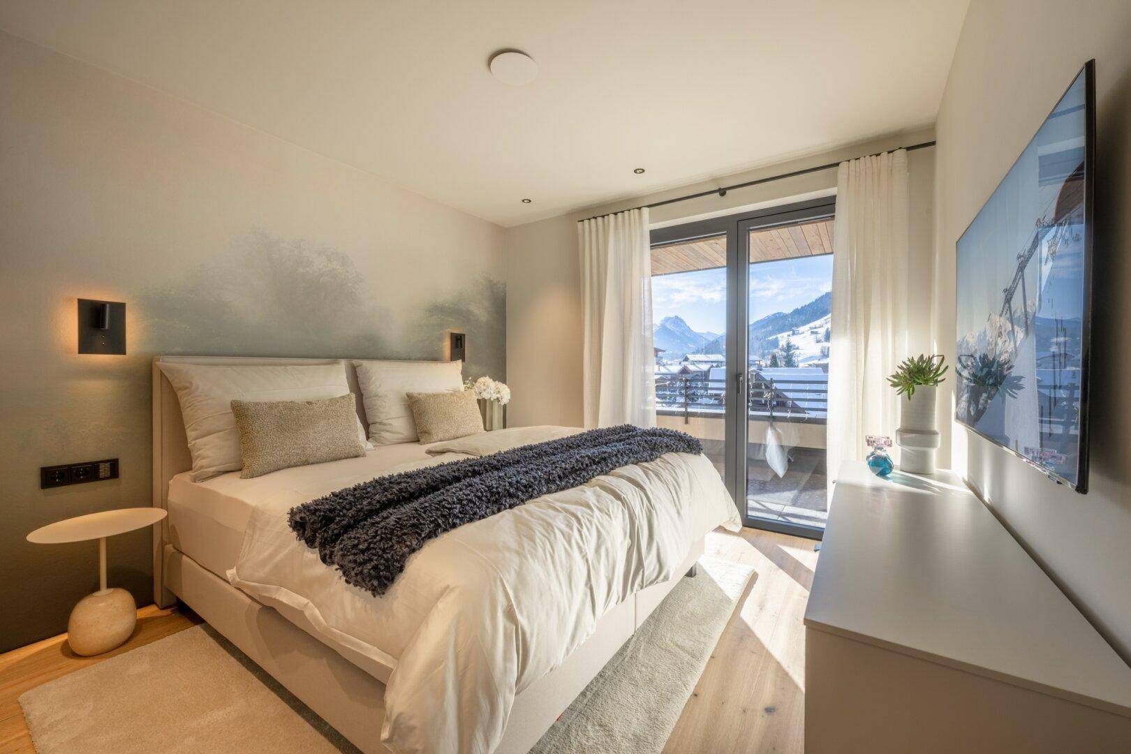 Helles Schlafzimmer mit Doppelbett, Fernseher und Balkonzugang mit Bergblick.