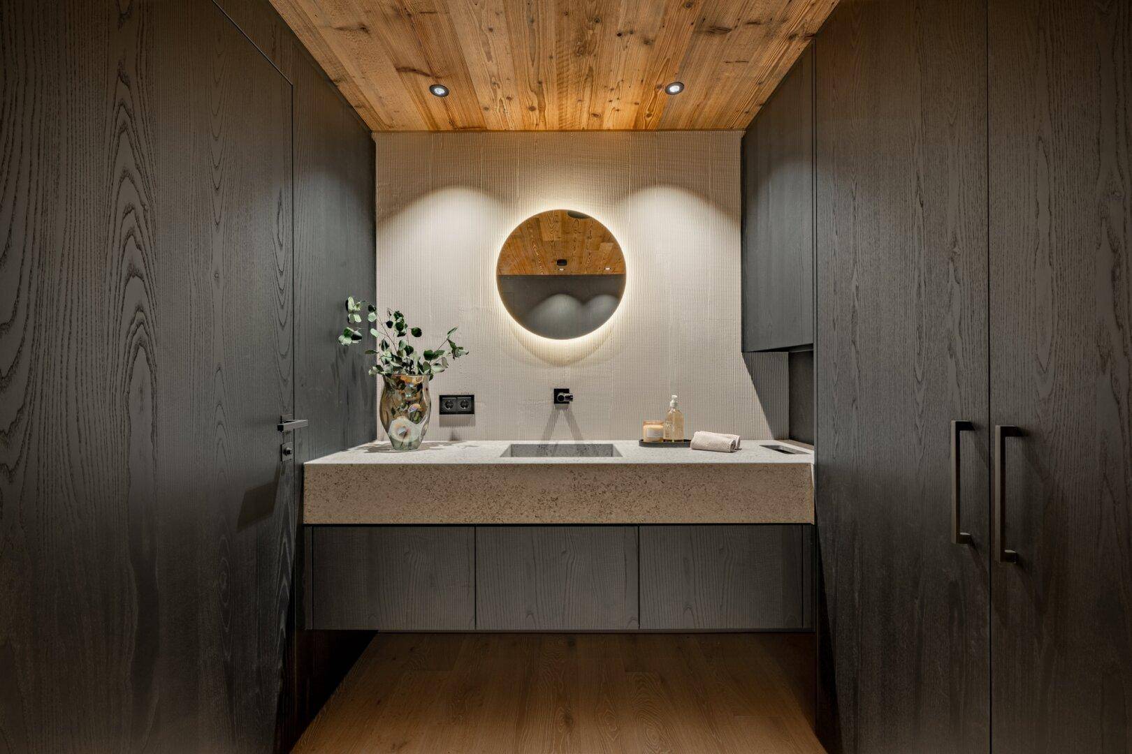 Modernes Gäste-WC mit rundem Spiegel, Waschbecken und dunklen Holzschränken.