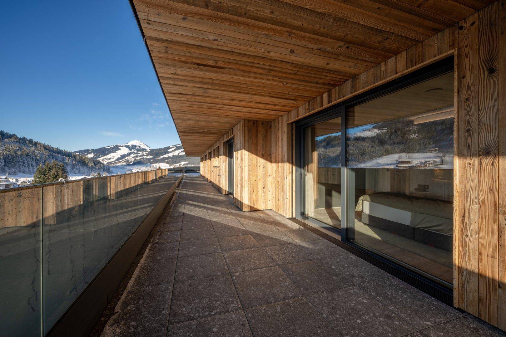 Langer Balkon mit Holzdach, Glasgeländer und weitem Blick auf die verschneite Berglandschaft.