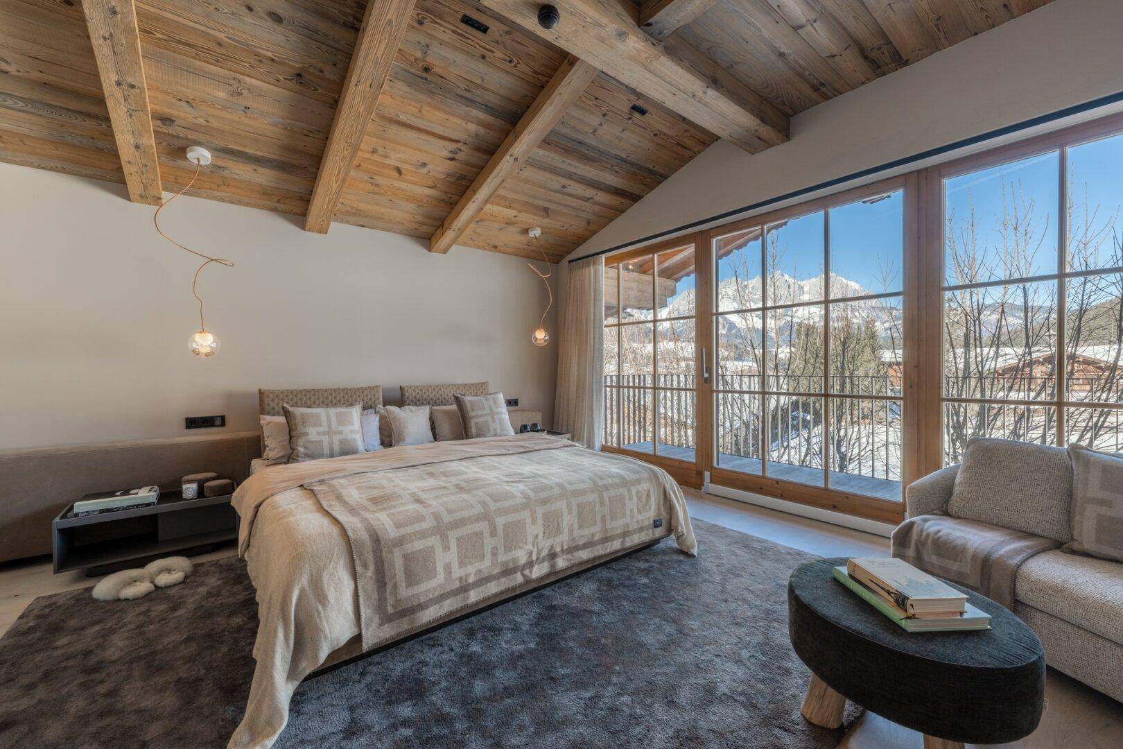 Helles Schlafzimmer mit Holzbalkendecke, großen Fenstern und direktem Blick auf die verschneiten Berge.