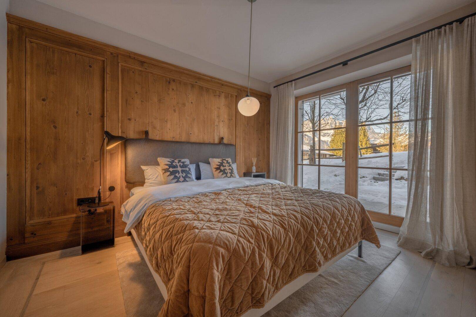Komfortables Schlafzimmer mit Holzwand, großen Fenstern und Zugang zum Balkon mit Bergblick.