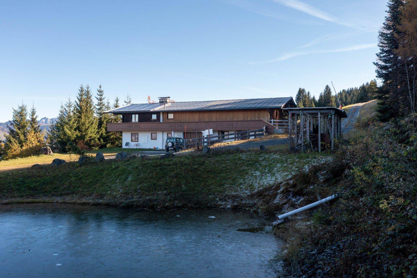 Haus am Hang mit Blick auf einen teilweise zugefrorenen Teich und die umliegende Berglandschaft.