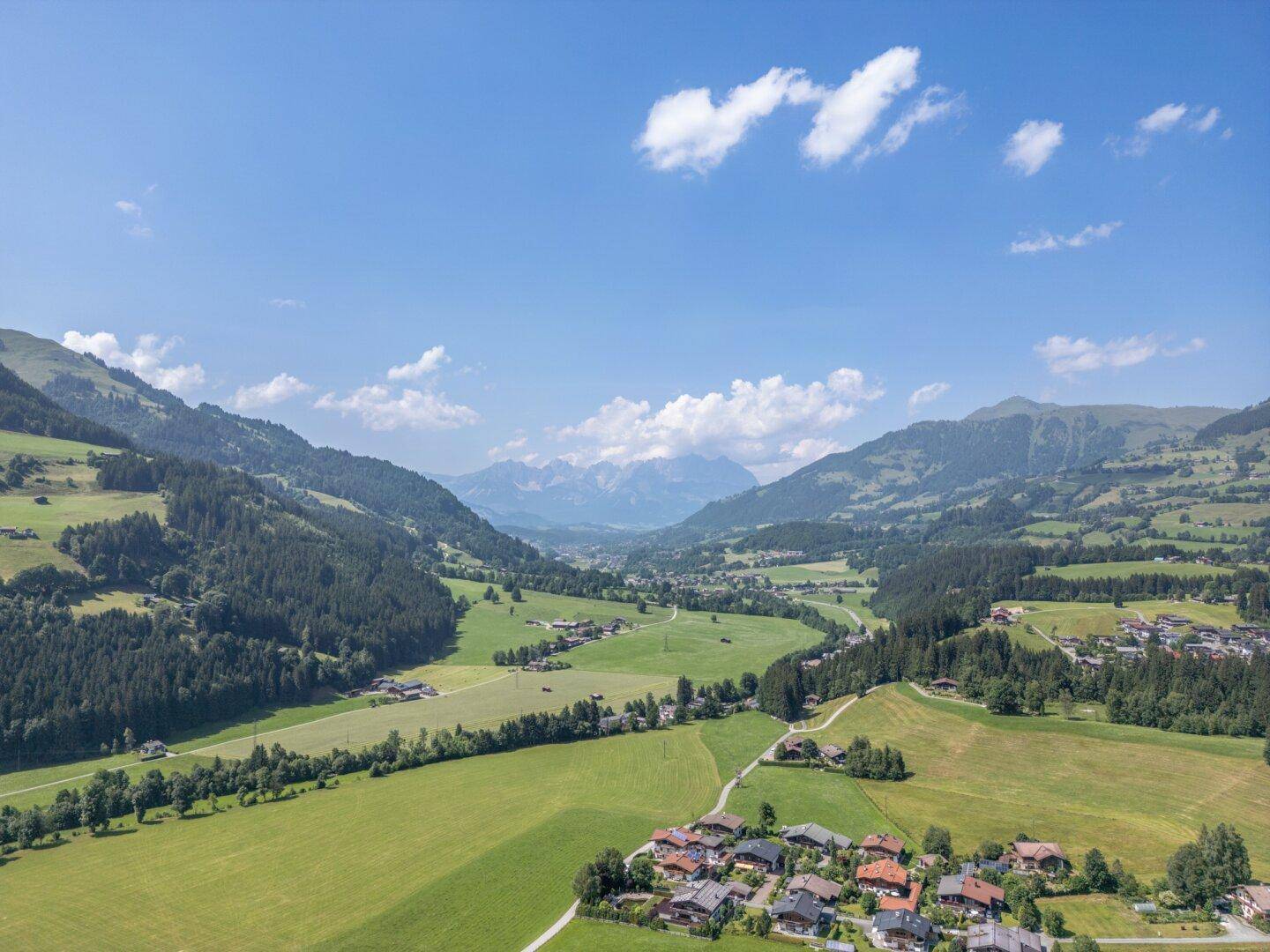 Weitläufige Landschaftsaufnahme eines grünen Tals mit Bergen und verstreuten Häusern unter blauem Himmel.