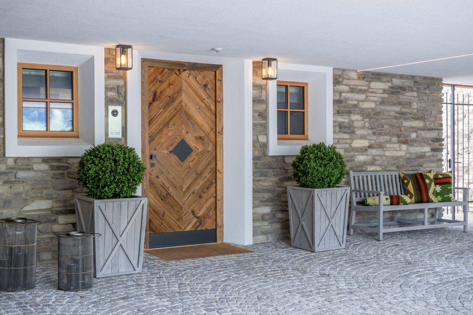 Eingangsbereich mit Holztür und Steinmauer, dekoriert mit Pflanzen und einer Sitzbank.