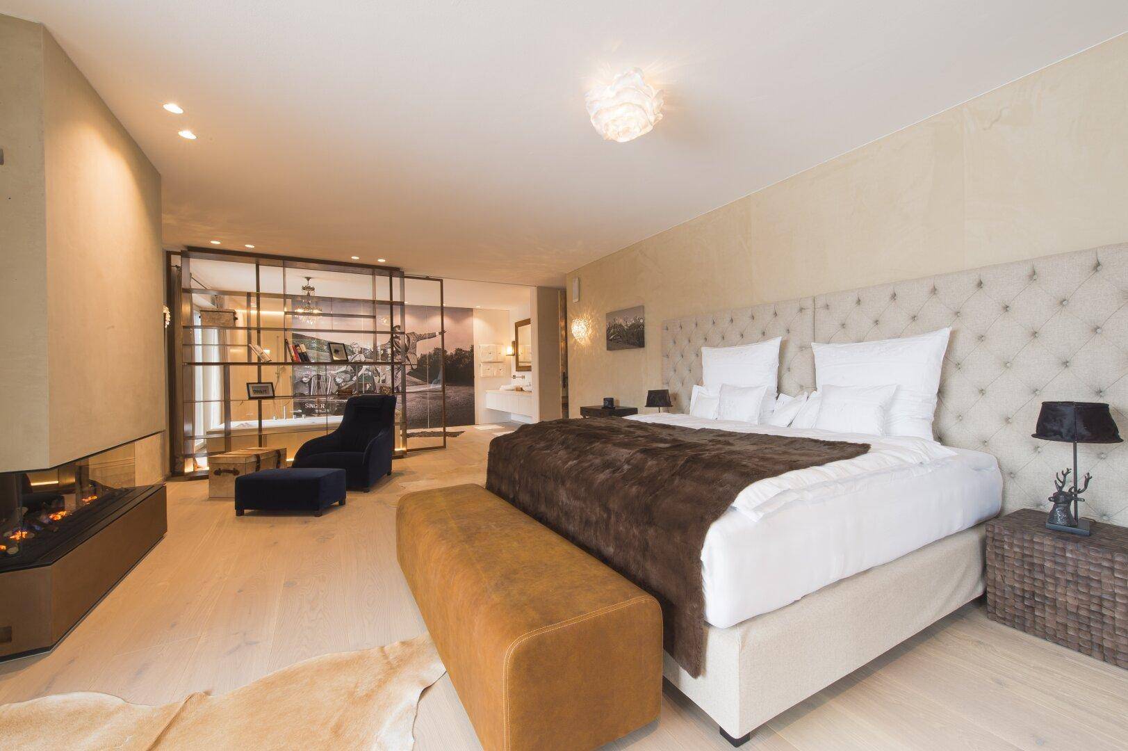 Elegantes Schlafzimmer mit großem Bett, Kamin und offenem Übergang zu einem stilvollen Sitzbereich.