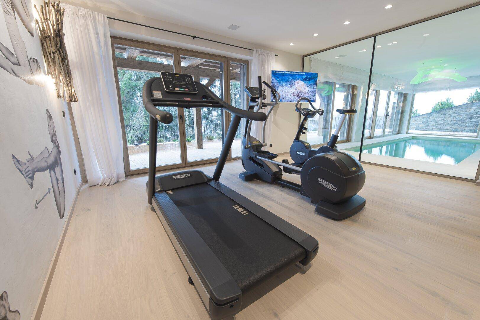 Fitnessraum mit Laufband, Crosstrainer und großen Fenstern mit Blick auf den Poolbereich.