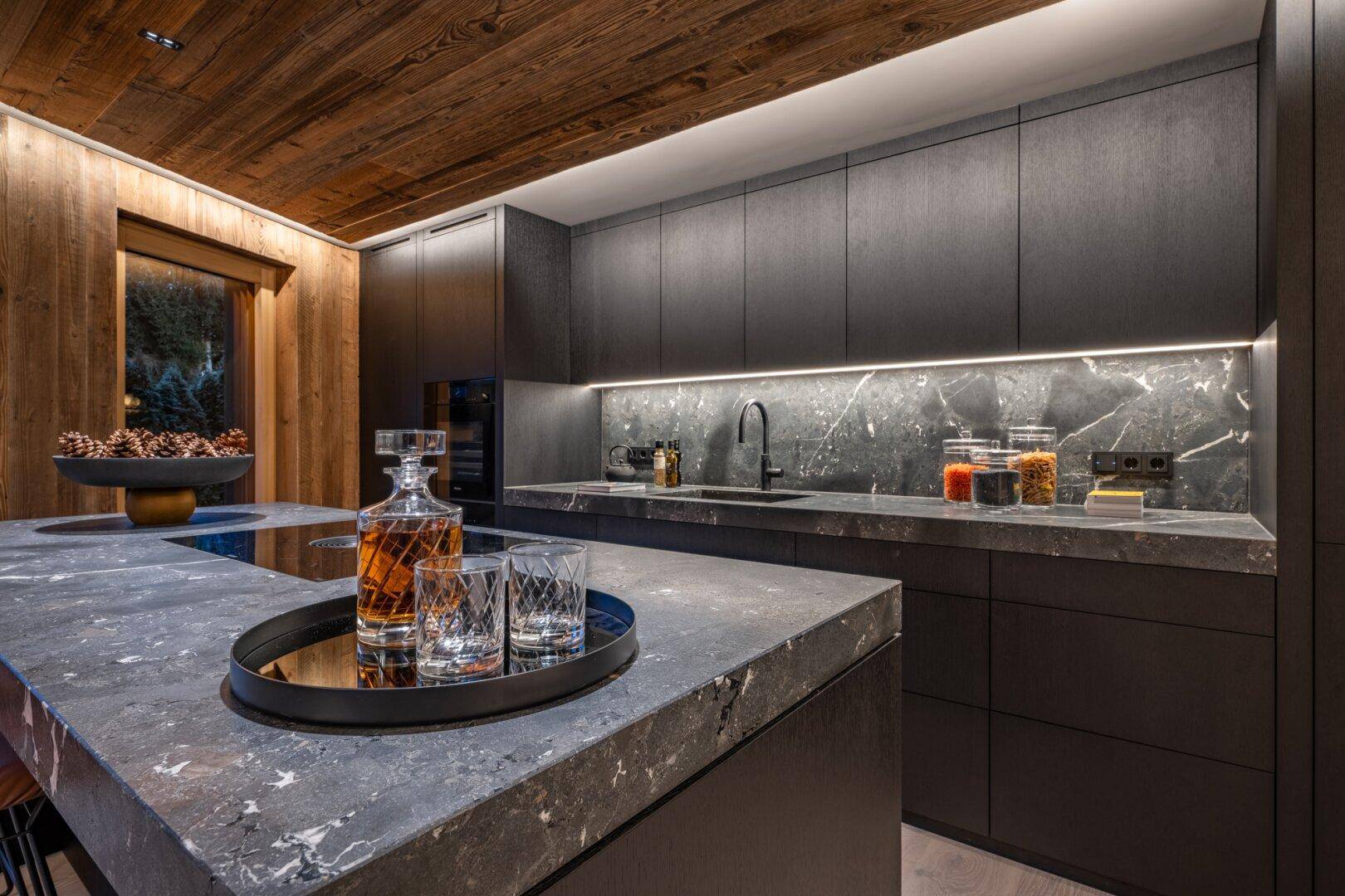Moderne Küche mit dunklen Schränken, Stein-Arbeitsplatten und Holzdecke, elegant beleuchtet.
