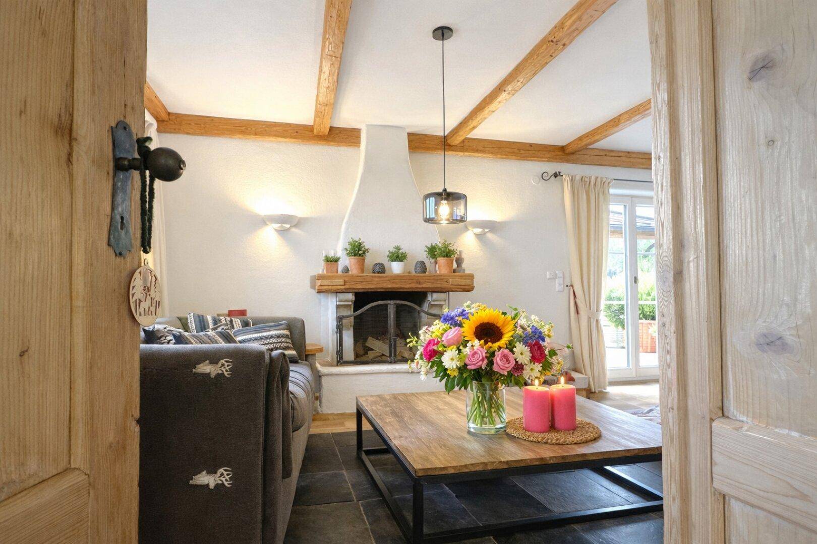 Gemütliches Wohnzimmer mit Kamin, Holzbalken und Zugang zu einem sonnigen Außenbereich.